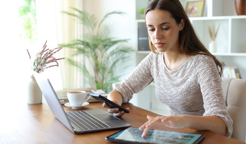 Image of a woman sitting at a desk with a phone, tablet, and laptop.