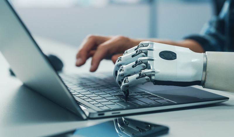 image of human and robotic hands typing at a laptop